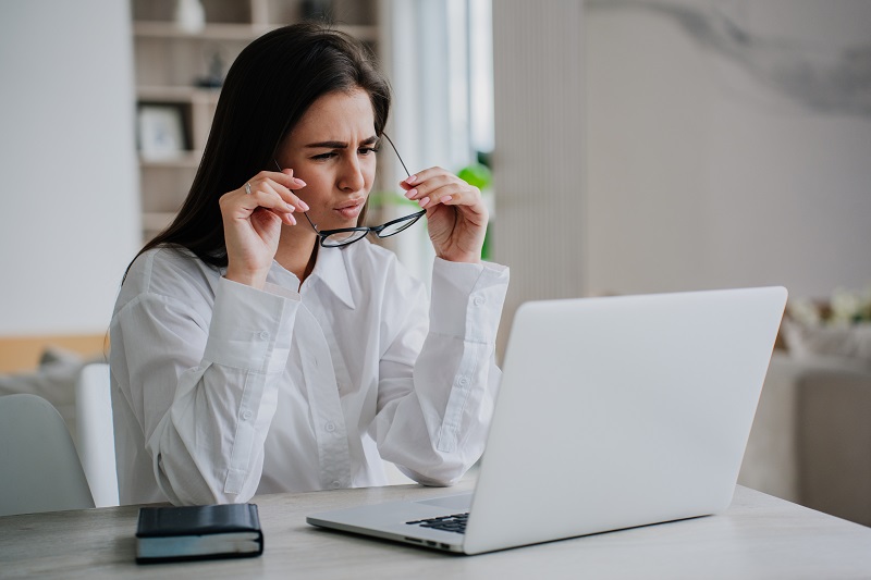 a person holding her glasses to her face while looking at a laptop.